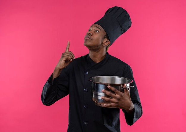 Free photo young surprised afro-american cook in chef uniform holds saucepan and points up isolated on pink wall