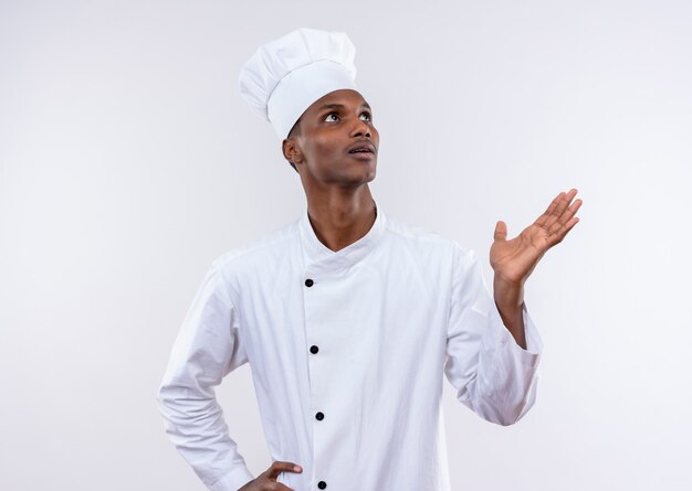Young surprised afro-american cook in chef uniform holdshand up and looks up isolated on white wall