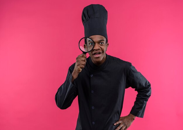 Young surprised afro-american cook in chef uniform looks through magnifying glass or loupe isolated on pink wall 