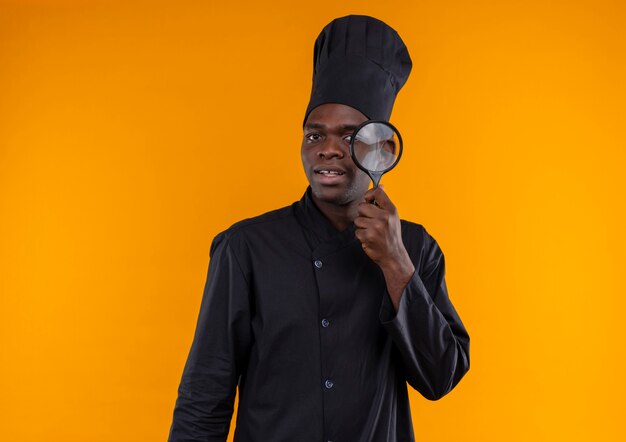 Young surprised afro-american cook in chef uniform looks through magnifying glass on orange  with copy space