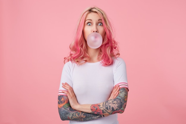 Free photo young surprised beautiful pink haired woman with tattooed hands, wears in white t-shirt, blows gum ball, looking to the left in surprise, stands.