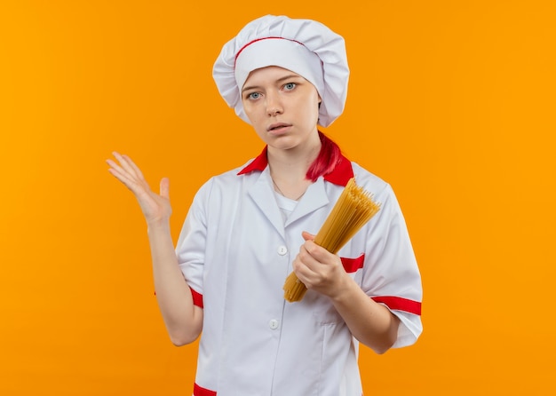 Il giovane cuoco unico femminile biondo sorpreso in uniforme del cuoco unico tiene il mazzo di spaghetti e punti a lato con la mano isolata sulla parete arancione