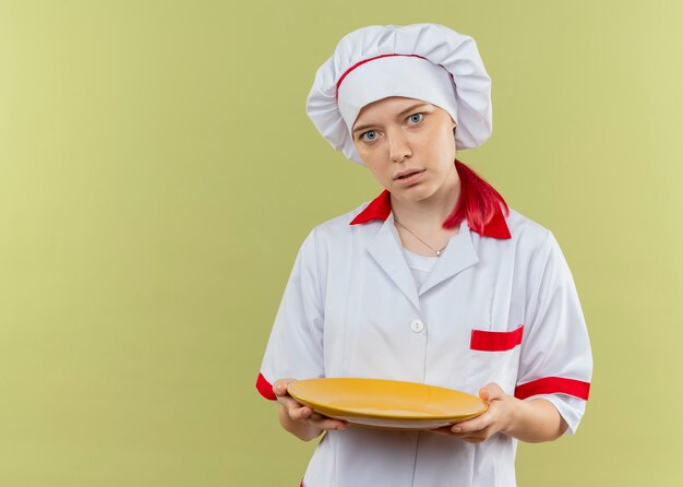 Молодая удивленная блондинка-шеф-повар в форме шеф-повара держит тарелку и выглядит изолированной на зеленой стене