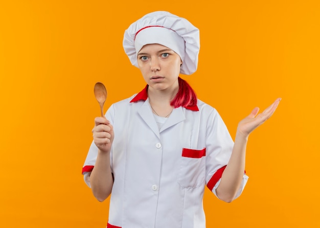 Young surprised blonde female chef in chef uniform holds spoon and holds hand open isolated on orange wall