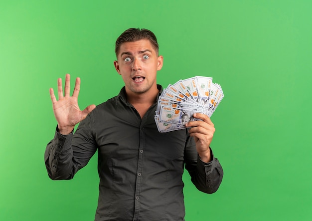 Young surprised blonde handsome man gestures five and holds money isolated on green space with copy space