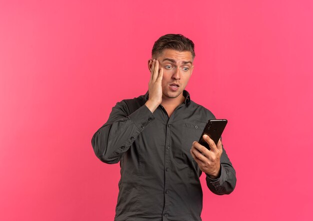 Young surprised blonde handsome man holds and looks at phone holding face isolated on pink background with copy space