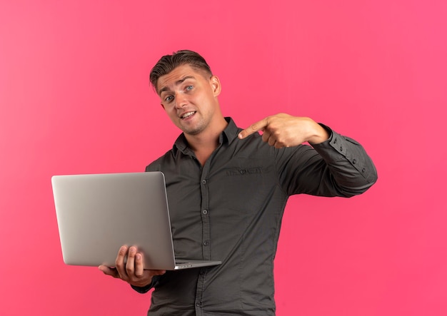 Young surprised blonde handsome man holds and points at laptop isolated on pink background with copy space