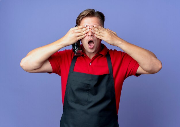 Young surprised blonde male barber in uniform closes eyes with hands holding hair clipper isolated on violet space with copy space