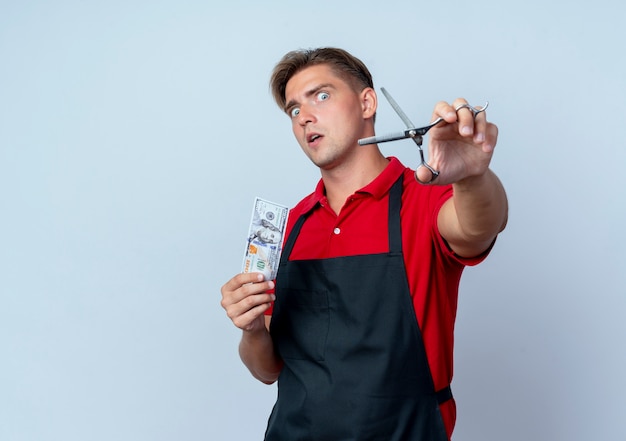 Young surprised blonde male barber in uniform holds hundred dollars bill and scissors isolated on white space with copy space