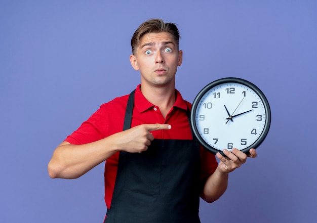 Young surprised blonde male barber in uniform holds and points at clock isolated on violet space with copy space