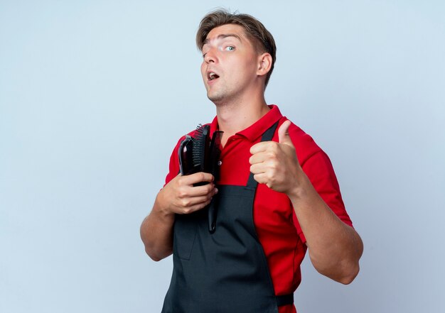 Young surprised blonde male barber in uniform thumbs up holding barber tools isolated on white space with copy space