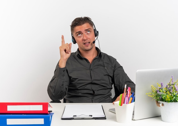 L'uomo giovane sorpreso dell'operaio di ufficio biondo sulle cuffie si siede alla scrivania con gli strumenti dell'ufficio utilizzando gli sguardi del computer portatile e indica in alto isolato su priorità bassa bianca con lo spazio della copia