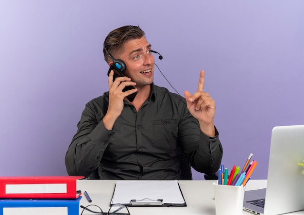 Young surprised blonde office worker man on headphones sits at desk with office tools using laptop talks on phone pointing up isolated on violet background with copy space