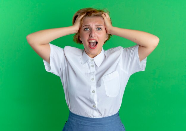 Young surprised blonde russian girl holds head looking at camera isolated on green background with copy space