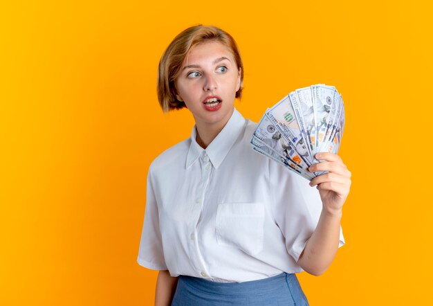 Young surprised blonde russian girl holds and looks at money isolated on orange background with copy space