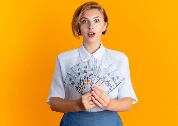 Young surprised blonde russian girl holds money isolated on orange background with copy space