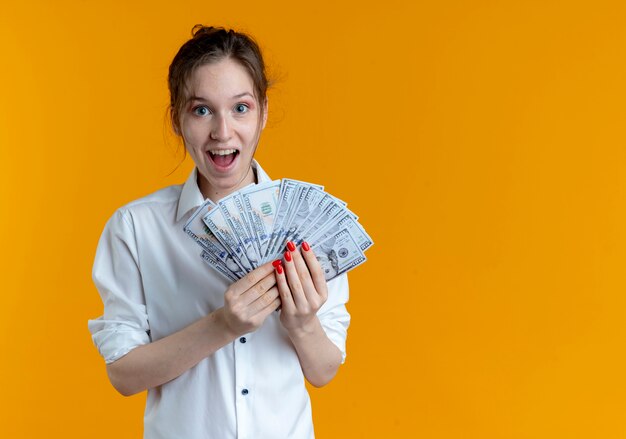 Young surprised blonde russian girl holds money isolated on orange space with copy space