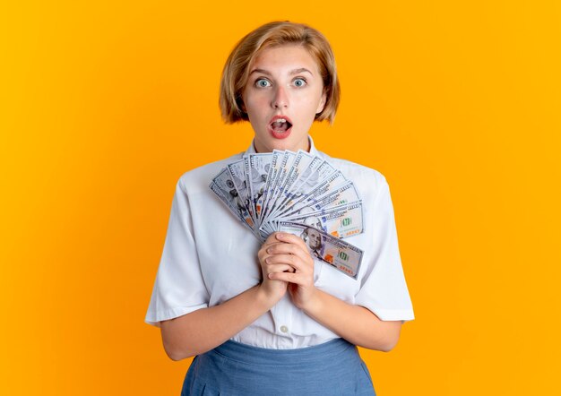 Young surprised blonde russian girl holds money looking at camera isolated on orange background with copy space