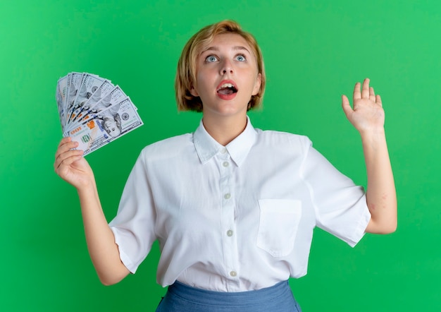 Young surprised blonde russian girl holds money with raised hand looking up isolated on green background with copy space