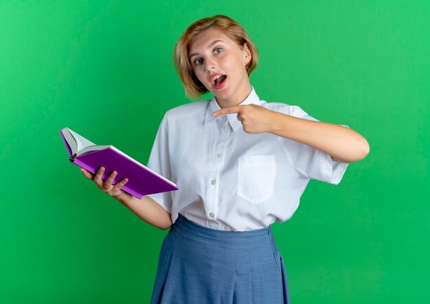 Молодая удивленная русская блондинка держит и указывает на книгу, изолированную на зеленом фоне с копией пространства
