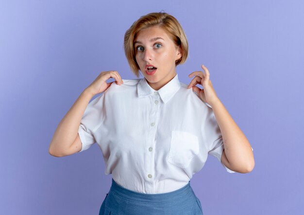 Young surprised blonde russian girl holds shirt looking at camera isolated on purple background with copy space