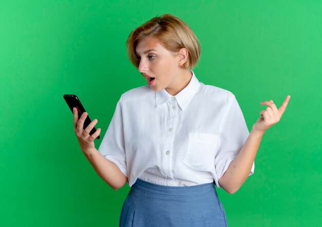 Young surprised blonde russian girl looks at phone isolated on green background with copy space