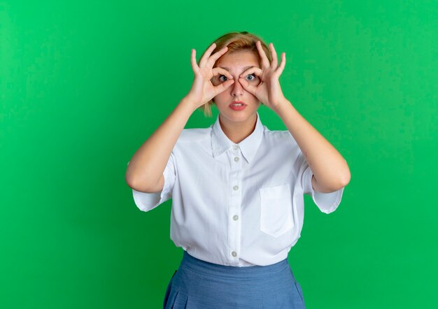 Young surprised blonde russian girl looks through fingers isolated on green background with copy space