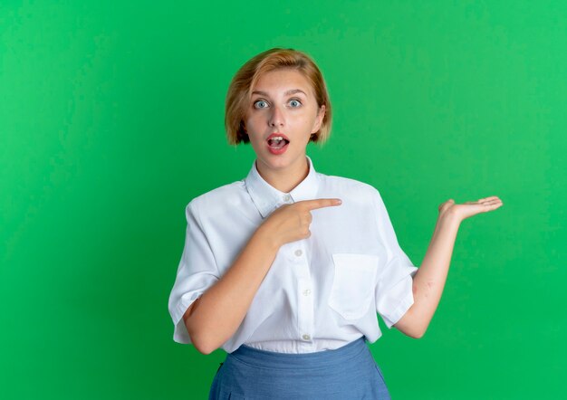 Young surprised blonde russian girl points at empty hand isolated on green background with copy space