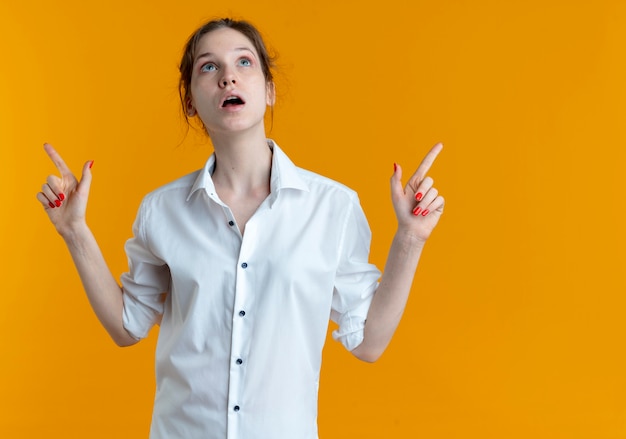 Young surprised blonde russian girl points and looks up isolated on orange space with copy space