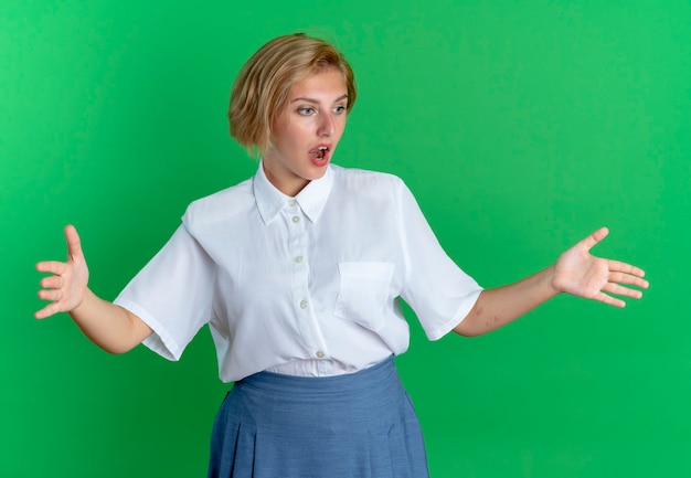 Young surprised blonde russian girl pretends to hold something and looks at it isolated on green background with copy space