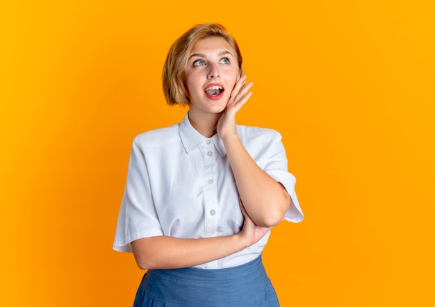 Young surprised blonde russian girl puts hand on chin looking at side isolated on orange background with copy space