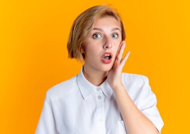 Young surprised blonde russian girl puts hand on face isolated on orange background with copy space