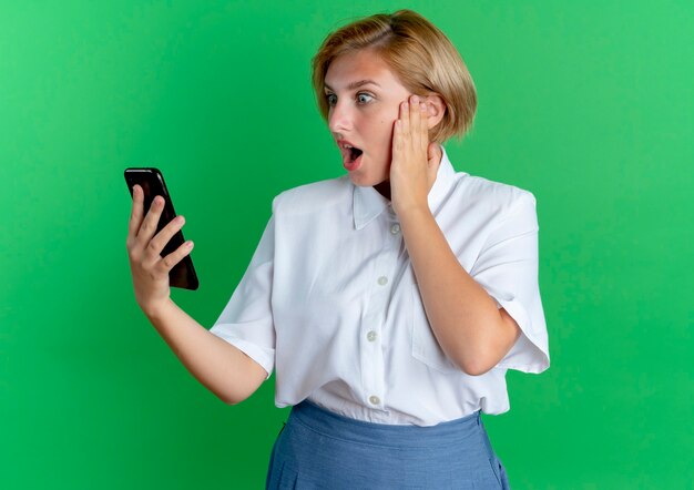 Foto gratuita giovane ragazza russa bionda sorpresa mette la mano sul viso guardando il telefono isolato su sfondo verde con spazio di copia