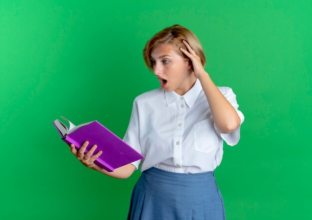 若い驚きの金髪のロシアの女の子は、コピースペースで緑の背景に分離された本を見て頭に手を置きます