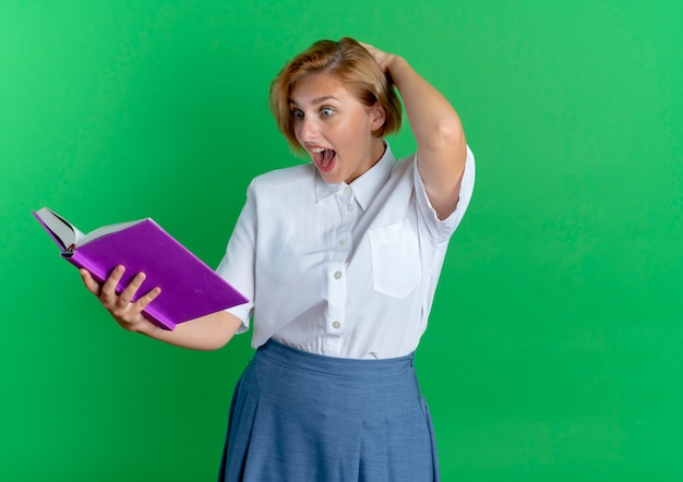 Free photo young surprised blonde russian girl puts hand on head behind looking at book