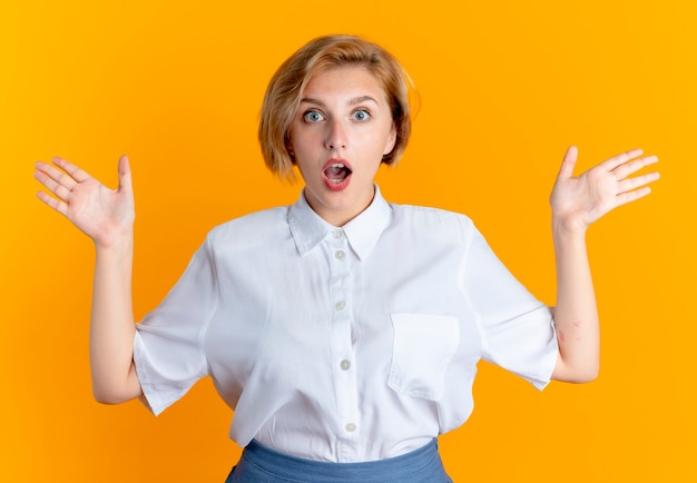 Young surprised blonde russian girl stands with raised hands isolated on orange background with copy space