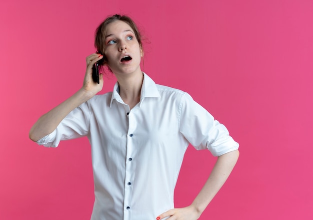 Young surprised blonde russian girl talks on phone looking at side isolated on pink space with copy space