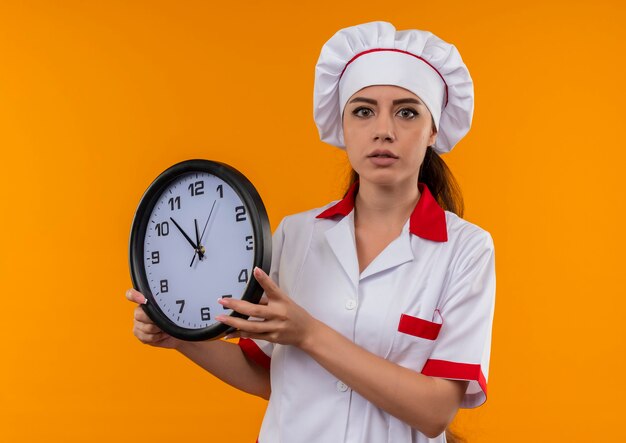シェフの制服を着た若い驚きの白人料理人の女の子は、コピースペースでオレンジ色の壁に分離された時計を保持します