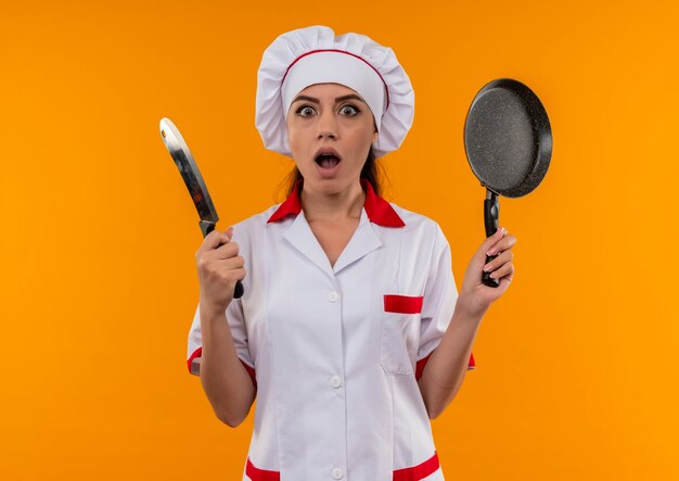 Молодая удивленная кавказская девушка-повар в униформе шеф-повара держит нож и сковороду, изолированные на оранжевой стене с копией пространства