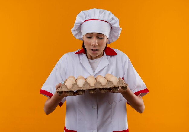 シェフの制服を着た若い驚きの白人料理人の女の子がコピースペースでオレンジ色の卵のバッチを保持し、見て