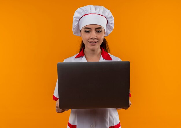 Young surprised caucasian cook girl in chef uniform holds and looks at laptop isolated on orange wall with copy space