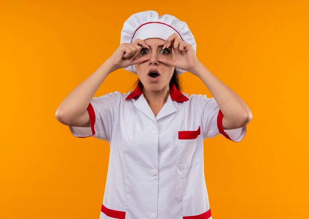 Free photo young surprised caucasian cook girl in chef uniform opens eyes with fingers isolated on orange wall with copy space