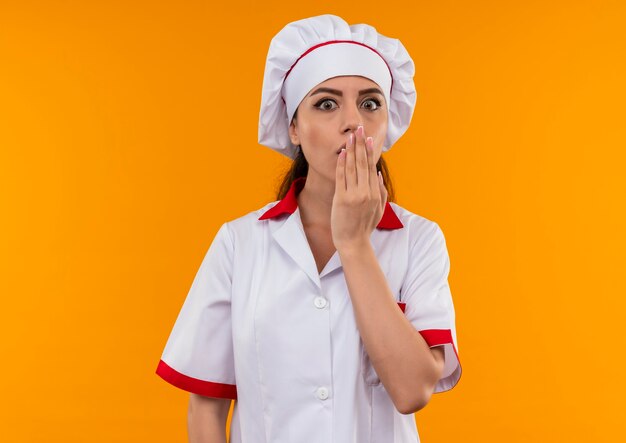 Young surprised caucasian cook girl in chef uniform puts hand on mouth isolated on orange wall with copy space