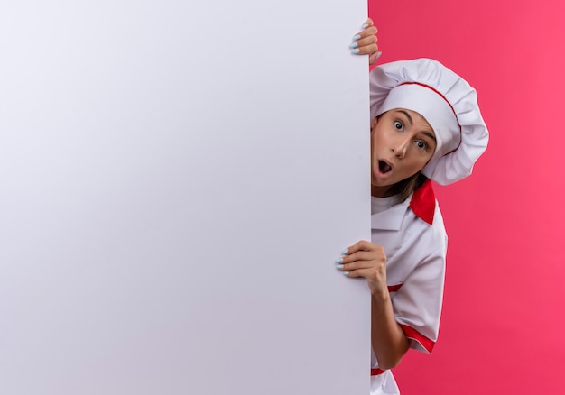 Free photo young surprised caucasian cook girl in chef uniform stands behind white wall on pink  with copy space