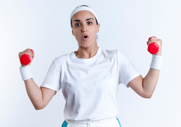 Young surprised caucasian sporty woman wearing headband and wristbands holds dumbbells isolated on white space with copy space