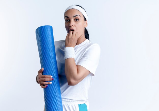 young surprised caucasian sporty woman wearing wristbands puts hand on mouth holding sports mat