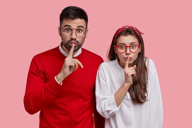 Free photo young surprised couple make silence sign, stand closely to each other, keep index fingers on lips, demonstrate shush gesture, ask not tell secret