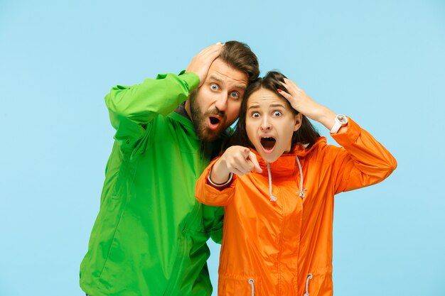 The young surprised couple pointing to camera and posing at studio in autumn jackets isolated on blue.