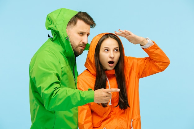 young surprised couple pointing to left and posing at studio in autumn jackets isolated on blue. Human negative emotions. Concept of the cold weather. Female and male fashion concepts
