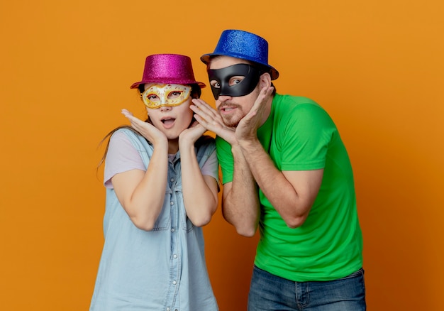 Young surprised couple wearing pink and blue hats put on masquerade eye masks putting hands on chin isolated on orange wall